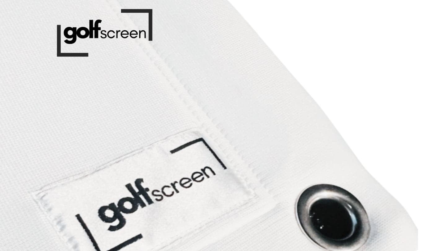 Corner of a finished white impact screen: a metal eyelet set through a folded, twin-stitched hem, with a woven golfscreen label sewn alongside it